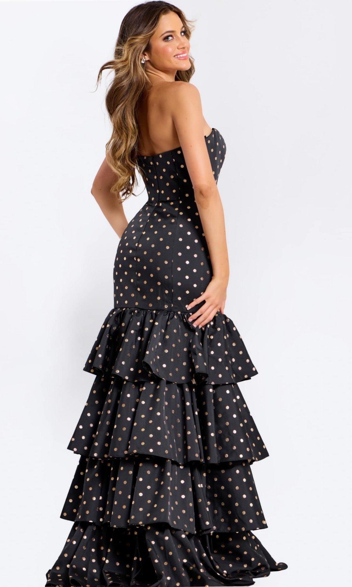 Woman modeling the Long Prom Dress: Jovani 49419—a strapless black gown with gold polka dots and a ruffled tiered skirt—posing sideways against a plain white background.